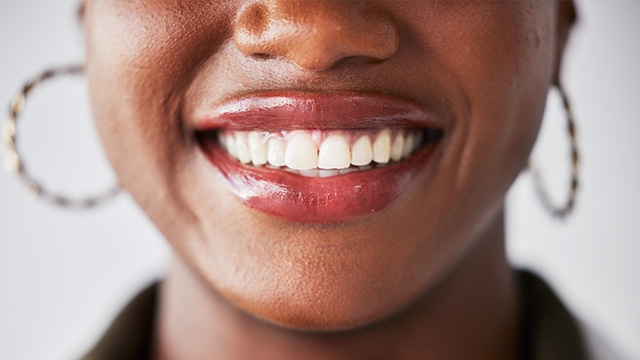 The image shows a person smiling at the camera with their mouth open, revealing teeth. They have dark skin and are wearing hoop earrings. The background is neutral and does not provide any additional context about the setting or occasion.
