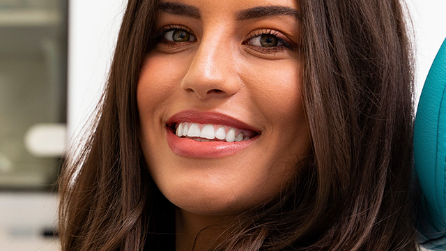 The image shows a smiling woman with short hair, wearing makeup and a black top, against a blurred background.