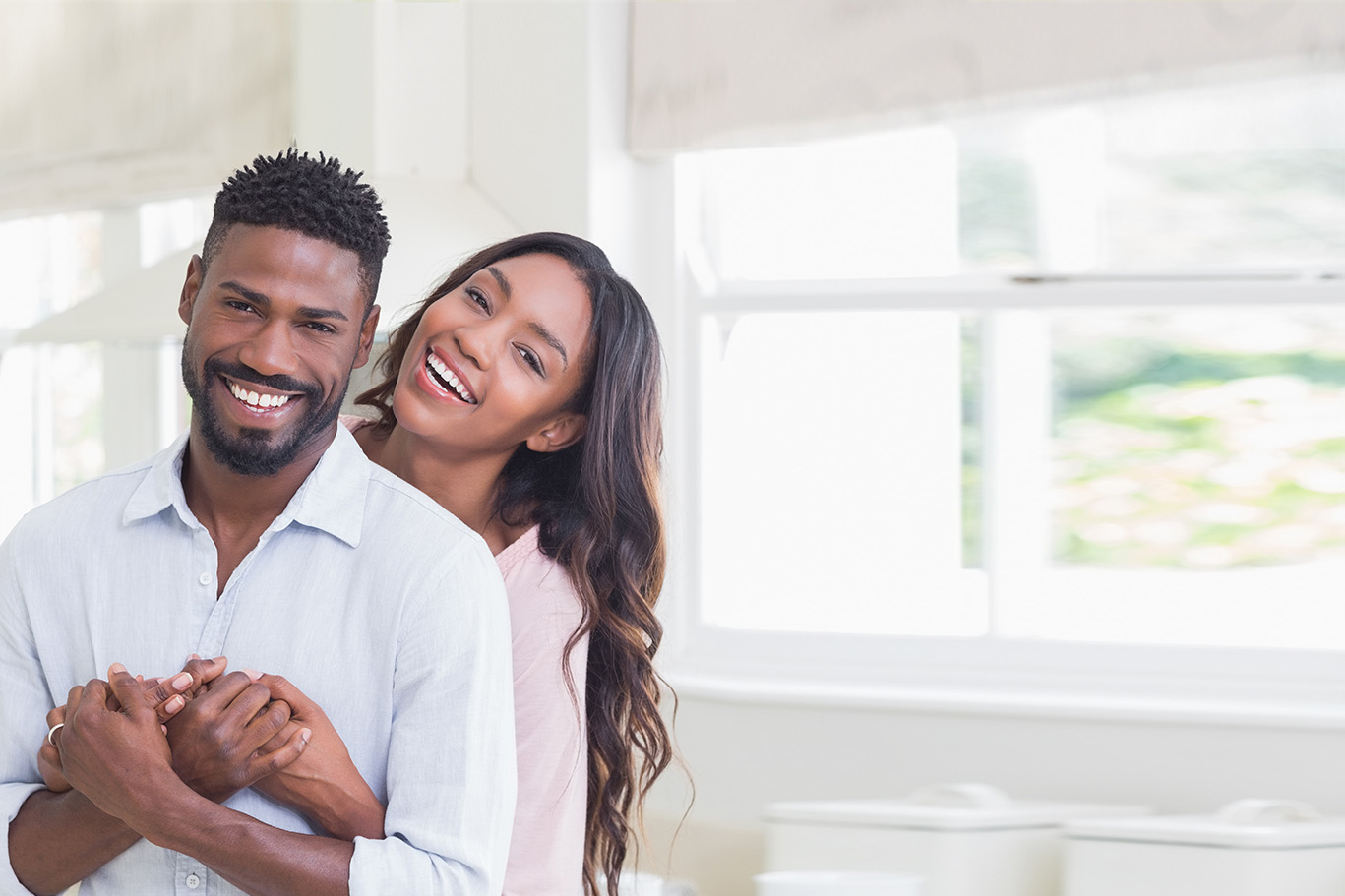 A man and a woman, both smiling, embrace in front of a window with blinds.